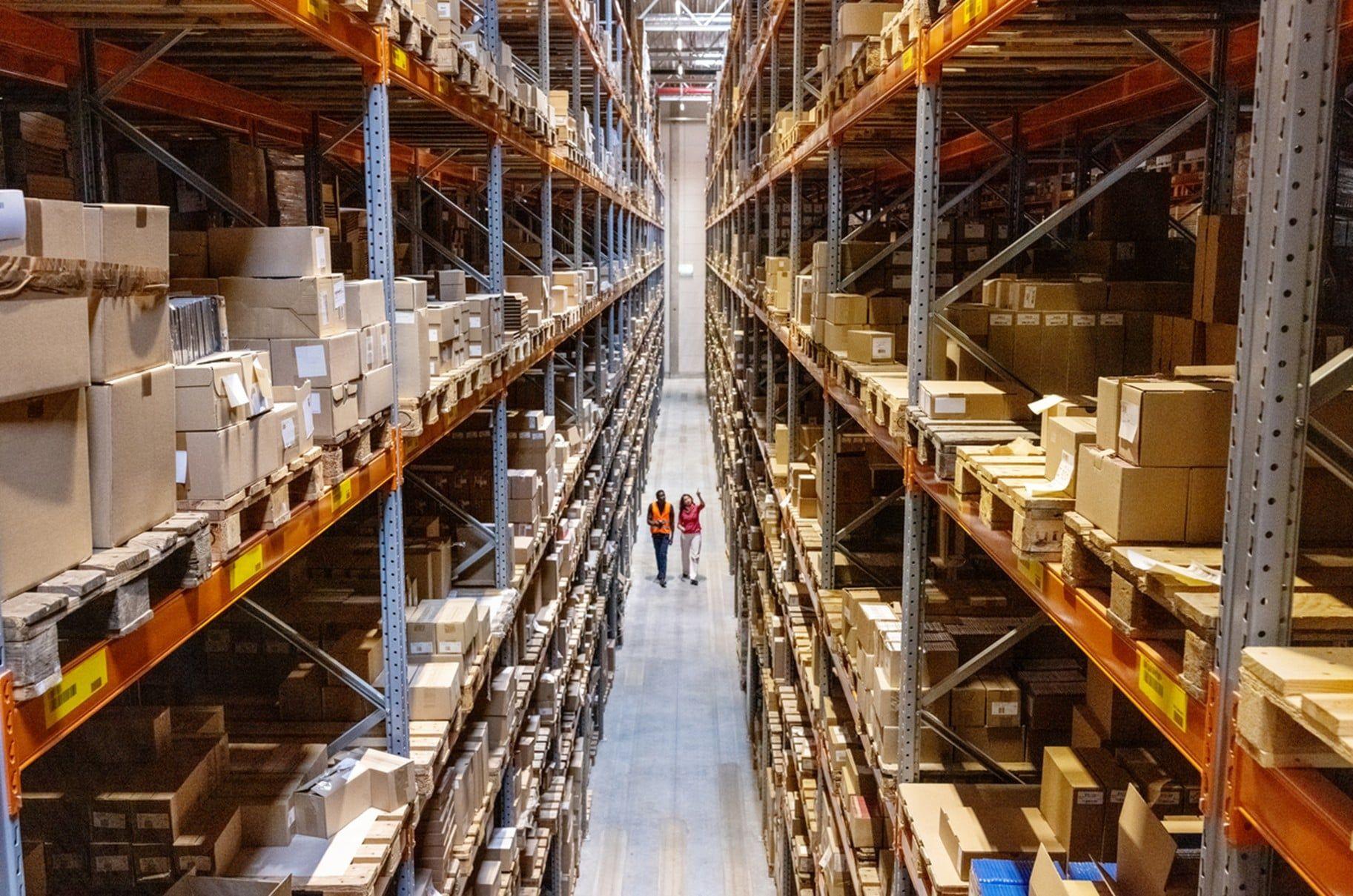 Two workers in high-visibility vests walking down an aisle between tall warehouse racking