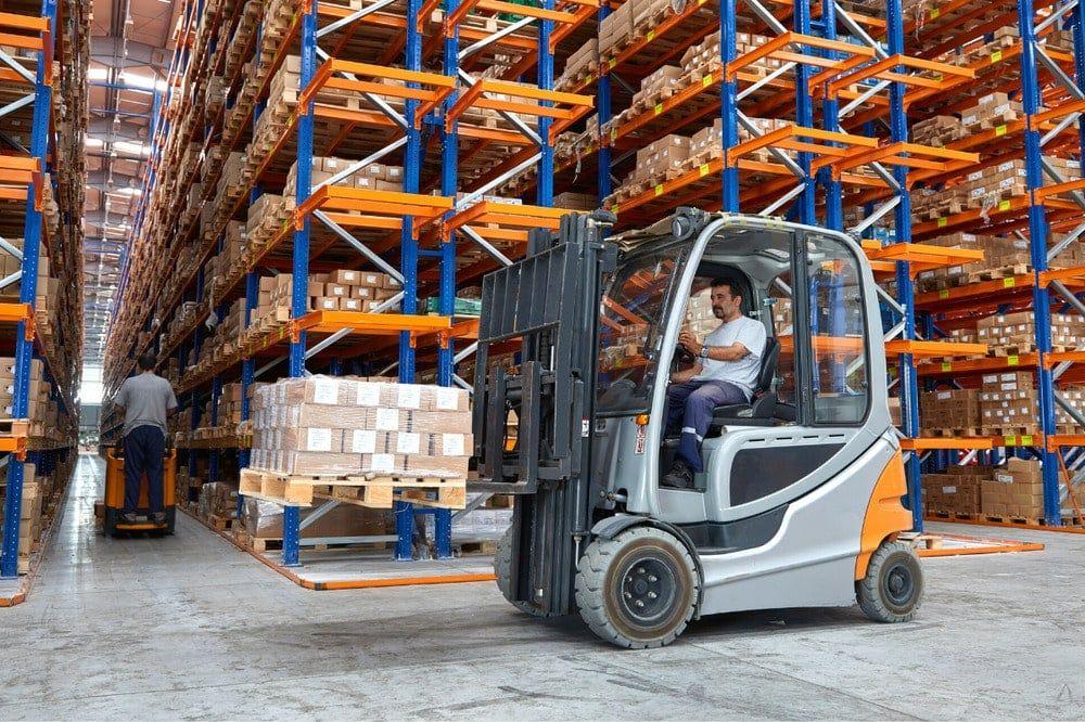Forklift driver in a warehouse aisle lined with tall blue and orange racking full of cartons
