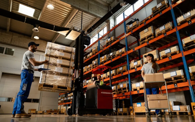 Warehouse workers moving wrapped pallets and cartons past orange racking stacked with crates