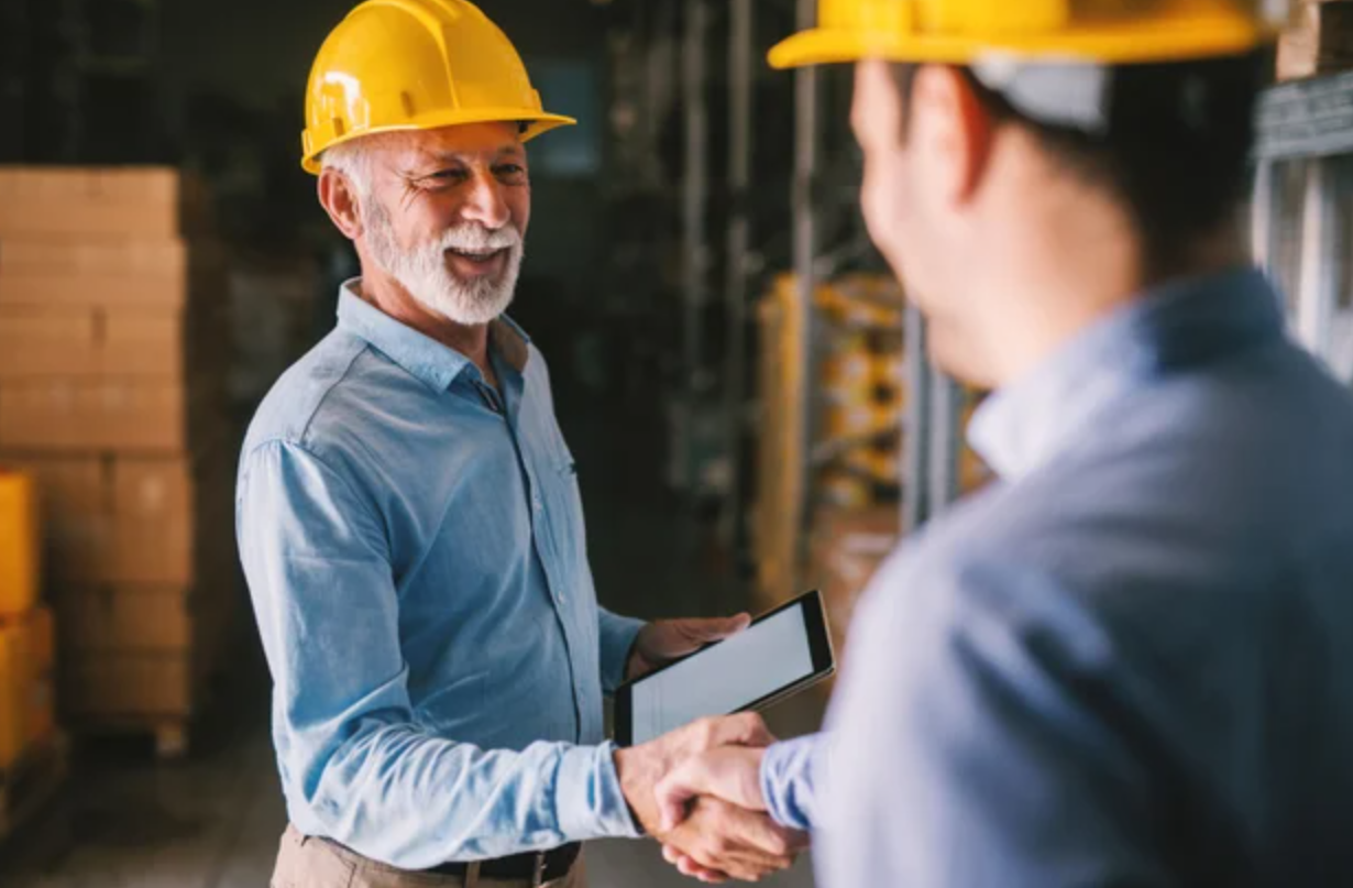 Two workers in hard hats shaking hands in a warehouse one holding a tablet