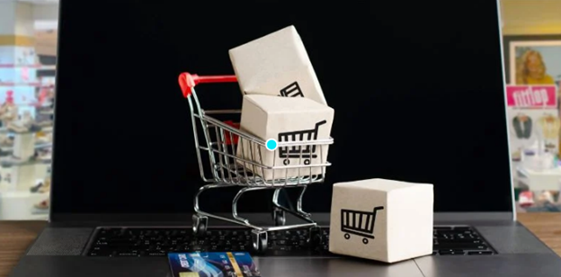 Miniature shopping cart holding small cardboard boxes on a laptop keyboard beside credit cards