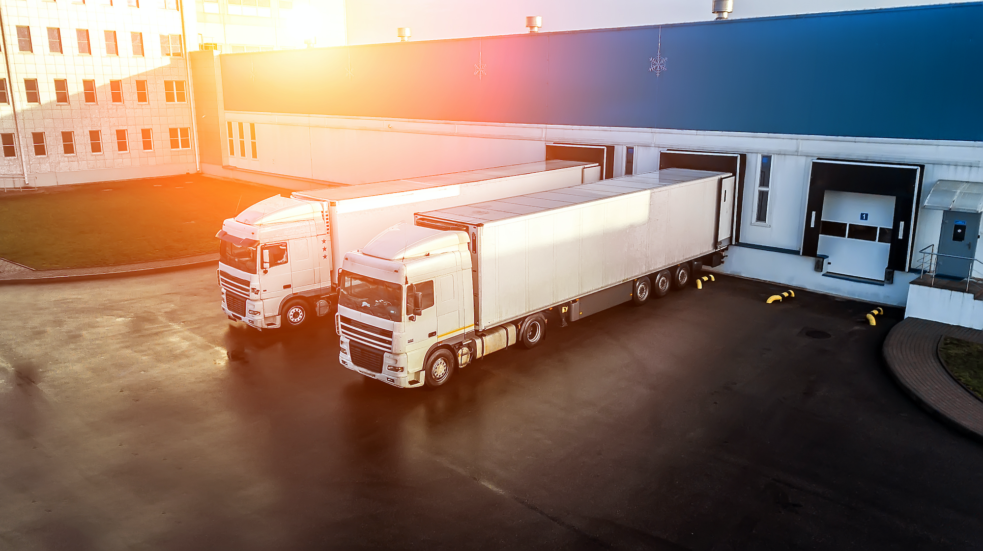 Two white semi-trailer trucks parked in a yard beside warehouse loading dock doors at sunrise